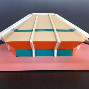 Op de afbeelding: Een kleurrijk, geometrisch model van een gebouw met een beige dak, oranje en blauwgroene muren en een roze basis. Witte verticale steunen en blauwgroene strepen accentueren het dak. Het model is een miniatuur architectonisch ontwerp.
