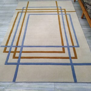 Puede incluir: Alfombra rectangular beige con diseño geométrico. La alfombra presenta cuadrados y rectángulos superpuestos en azul y marrón, creando un patrón abstracto moderno. La alfombra está colocada sobre un suelo de color claro. Medidas en centímetros.