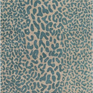 Op de afbeelding: Een vierkant tapijt met een herhalend dierenpatroon in blauwgroen op een neutrale beige achtergrond. Het ontwerp bestaat uit onregelmatige, organische vormen die een luipaardvlekkenmotief nabootsen en het gehele oppervlak van het textiel bedekken.