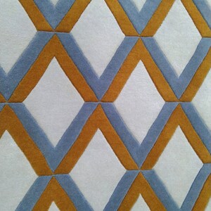 May include: Close-up of a geometric patterned rug. The rug features a repeating diamond design with alternating colours. The diamonds are outlined in a mustard yellow colour, with the interior filled with a light blue colour. The background is white.