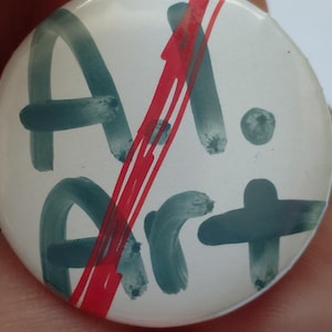 May include: A round white pin-back button featuring dark green hand-painted lettering that reads AI Art. A bold, bright red diagonal slash mark crosses through the center of the text, obscuring parts of the letters while creating a striking contrast.