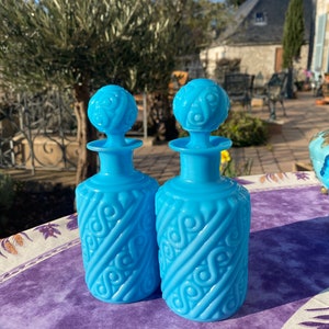 Puede incluir: Dos botellas de vidrio azul con tapones ornamentados y un diseño decorativo en relieve. Las botellas están sobre un mantel floral morado y blanco.