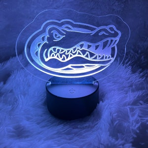 May include: A clear acrylic LED night light featuring a detailed outline of an alligator head within a rounded border. The light emits a cool blue glow and sits on a black circular base with a power symbol.