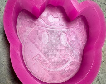 May include: Pink silicone mold shaped like a smiling face wearing a cowboy hat. The mold is designed for making cookies, fondant, or other edible treats.