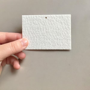 May include: A rectangular white felt tag with a small hole at the top. The tag has a textured surface and is held by a hand against a neutral gray background. The tag is blank and ready for use.