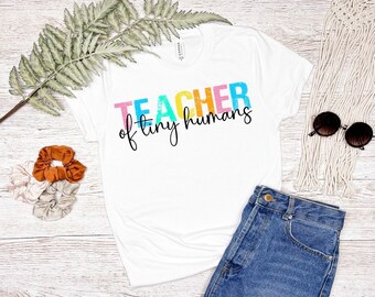 May include: White t-shirt with the text "TEACHER of tiny humans" in rainbow colors. The shirt is on a wooden surface with a pair of sunglasses, a denim skirt, and hair scrunchies.