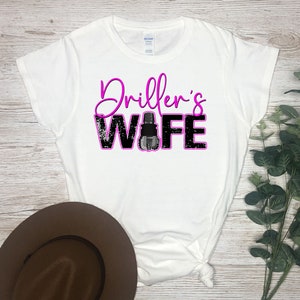 May include: White t-shirt with the text "Driller's Wife" in pink and black lettering. The word "Wife" has a black and gray drill bit graphic.