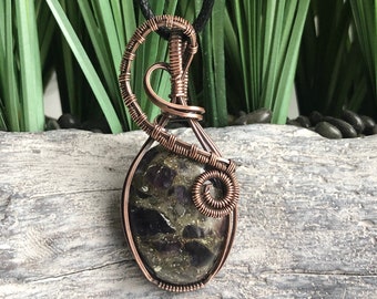 May include: A copper wire wrapped pendant featuring a green and black stone with a swirl design. The pendant is hanging from a black cord.