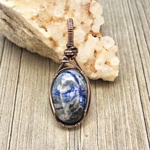 Puede incluir: Un colgante envuelto en alambre de cobre con una piedra ovalada azul y blanca. El colgante está sentado sobre un lecho de cristales blancos.