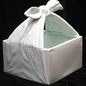 May include: White ceramic planter shaped like a box with a fabric-like lid tied with a bow. The planter is designed to look like a wrapped gift.