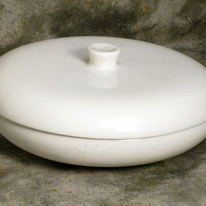 May include: A white, oval-shaped ceramic serving dish with a matching lid. The lid has a small, rounded handle. The dish and lid are smooth and glossy, with a simple, elegant design. The dish is suitable for serving food.