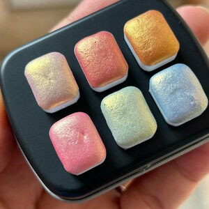 K&ouml;nnte beinhalten: Eine schwarze Metall-Aquarellfarbpalette mit sechs rechteckigen Pfannen mit schimmernden, irisierenden Farben. Die Farben umfassen Rosa, Rot, Gold, Gr&uuml;n und Blau, jede mit einem perlmuttartigen Finish. Die Palette wird in einer Hand gehalten.