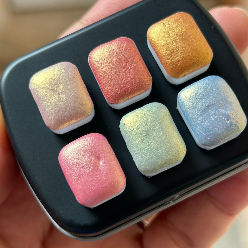 K&ouml;nnte beinhalten: Eine schwarze Metall-Aquarellfarbpalette mit sechs rechteckigen Pfannen mit schimmernden, irisierenden Farben. Die Farben umfassen Rosa, Rot, Gold, Gr&uuml;n und Blau, jede mit einem perlmuttartigen Finish. Die Palette wird in einer Hand gehalten.