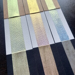 K&ouml;nnte beinhalten: Nahaufnahme einer Farbpalette mit metallischen Farbmustern. Die Muster sind auf Papierstreifen in verschiedenen Farben angeordnet, darunter Gold, Silber und Blau. Die Farbe hat ein schimmerndes, strukturiertes Aussehen, und der Hintergrund ist dunkel.