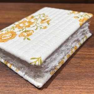 May include: A rectangular fabric-covered notebook featuring a cream-coloured quilted exterior with a yellow floral pattern. The edges of the pages are frayed and textured, showing a white, fibrous appearance between the stitched fabric covers.