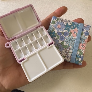 Puede incluir: Una paleta de acuarela de plástico rosa con 15 pocillos de pintura y una funda de tela floral. La funda tiene un patrón floral azul y blanco con una cinta azul claro.