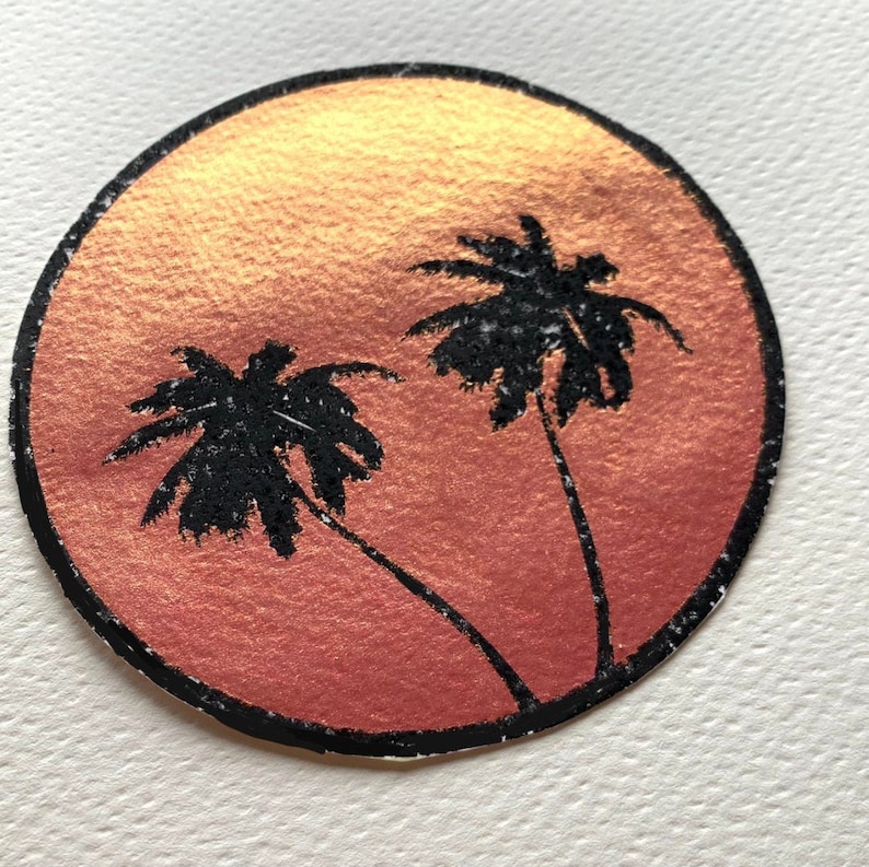 K&ouml;nnte beinhalten: Ein runder Aufkleber mit einer Sonnenuntergangsszene mit zwei schwarzen Palmen vor einem Farbverlauf aus Orange und Gold. Der Aufkleber hat einen schwarzen Rand und befindet sich auf einer wei&szlig;en Oberfl&auml;che.