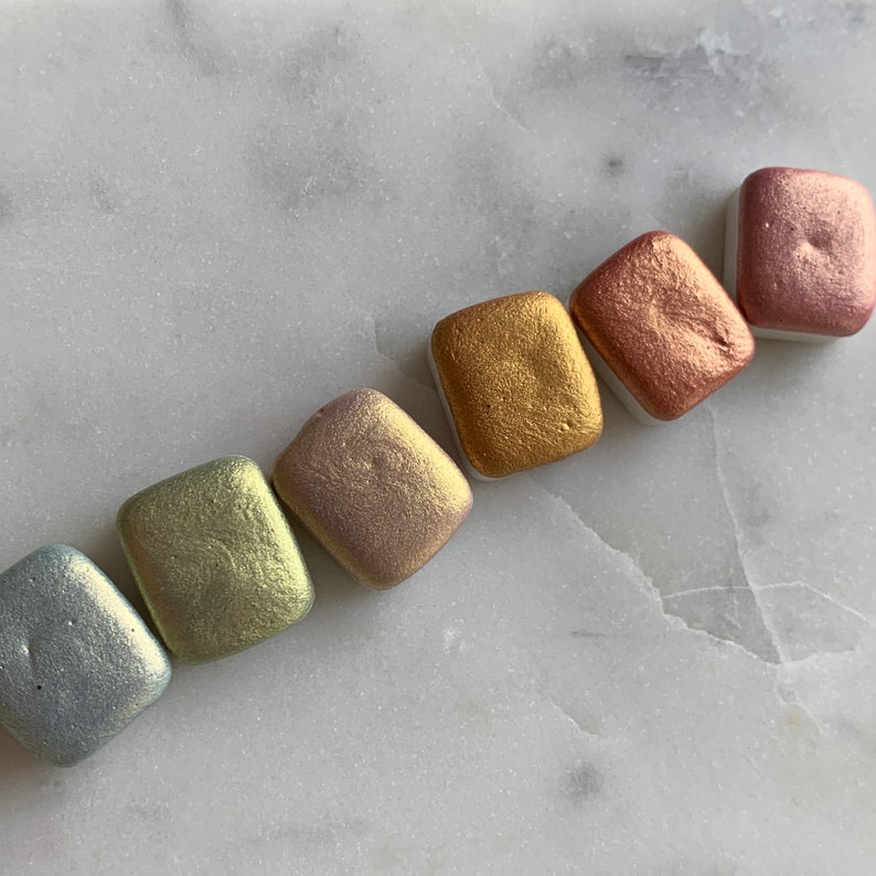 K&ouml;nnte beinhalten: Eine Sammlung von sieben quadratischen Aquarellfarben in verschiedenen Metallic-Farben, darunter Silber, Gold und Ros&eacute;. Die Farben sind diagonal auf einer wei&szlig;en Marmoroberfl&auml;che angeordnet und zeigen ihre schimmernden Pigmente.