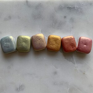 K&ouml;nnte beinhalten: Sechs metallische Aquarellfarben in einer Reihe, jede in einer anderen Farbe. Die Farben umfassen Silber, Hellgr&uuml;n, Ros&eacute;gold, Gold, Kupfer und Rosa. Die Pfannen sind rechteckig mit abgerundeten Ecken.