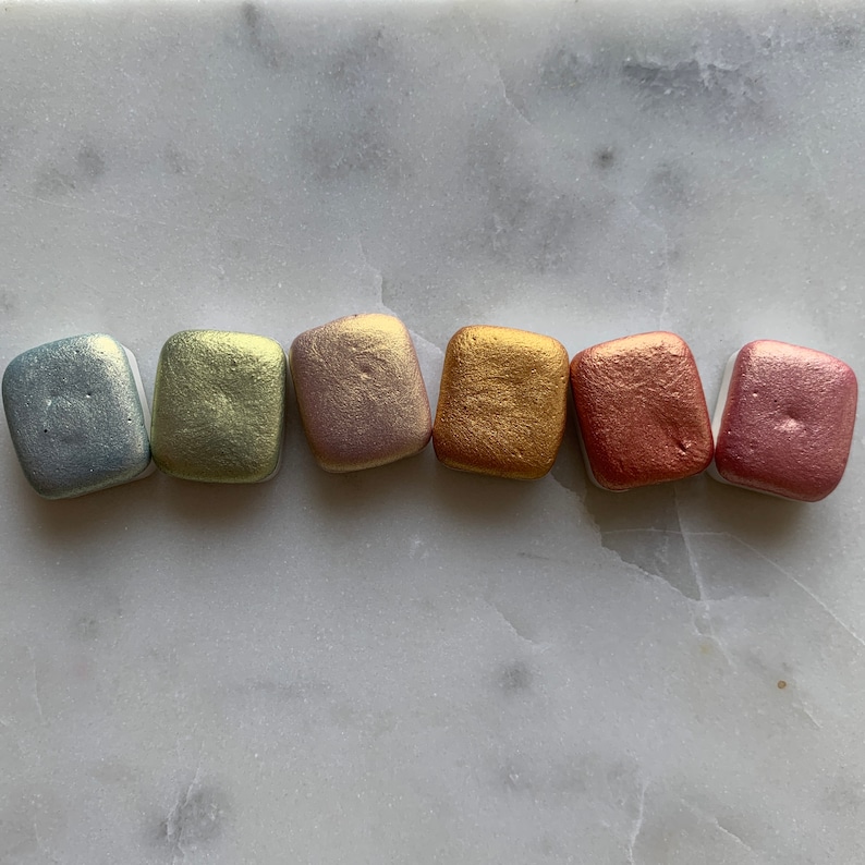 K&ouml;nnte beinhalten: Sechs metallische Aquarellfarben in einer Reihe, jede in einer anderen Farbe. Die Farben umfassen Silber, Hellgr&uuml;n, Ros&eacute;gold, Gold, Kupfer und Rosa. Die Pfannen sind rechteckig mit abgerundeten Ecken.