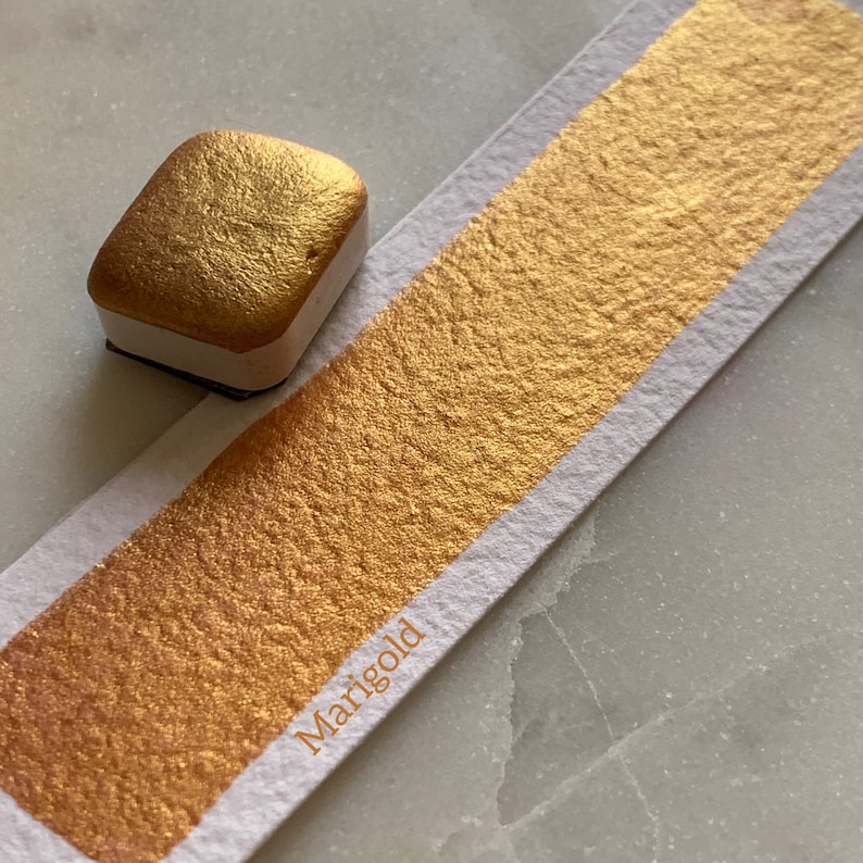 K&ouml;nnte beinhalten: Ein quadratischer Aquarellfarbkasten und ein Farbmuster. Die Farbe ist ein metallisches Gold, beschriftet mit "Marigold". Das Farbmuster ist ein rechteckiger Streifen Farbe auf wei&szlig;em Papier. Der Farbkasten ist goldfarben mit wei&szlig;er Basis.