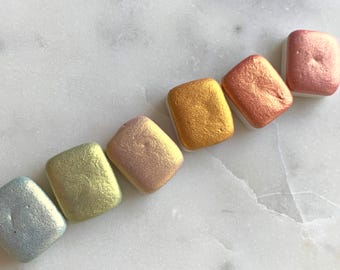 May include: Six square-shaped pans of metallic watercolor paint in shades of blue, green, pink, gold, copper, and silver.