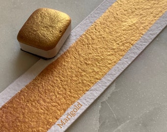 May include: A small, square pan of metallic gold watercolor paint. The paint is labeled "Marigold" and has a textured, metallic finish.