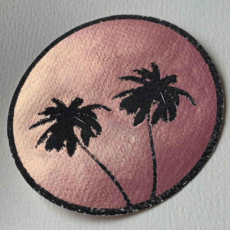 K&ouml;nnte beinhalten: Ein runder Aufkleber mit einem rosa Hintergrund mit Metallic-Effekt. Zwei schwarze Palmensilhouetten sind vor dem Rosa platziert. Der Aufkleber hat einen schwarzen Rand und eine strukturierte Oberfl&auml;che.