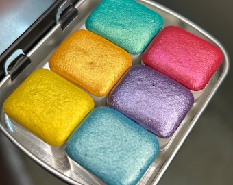 Puede incluir: Una lata de metal con seis bandejas de pintura de acuarela. Las bandejas están llenas de pinturas metálicas en tonos turquesa, dorado, rosa, morado y azul.