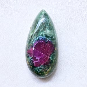Peut inclure: Une pierre précieuse en forme de goutte d'eau avec un centre rouge profond et vibrant et un extérieur vert moucheté. La surface de la pierre a un aspect légèrement texturé, avec des reflets bleus et violets.