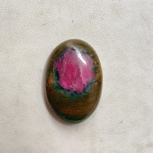 May include: An oval gemstone featuring a bright pink core, framed by green and brown hues. The polished stone has a smooth surface and a natural, earthy look. Suitable for jewellery or decorative purposes, this stone measures approximately 3 cm in length.