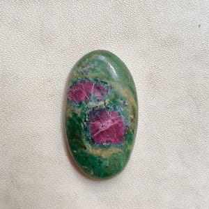 May include: Oval-shaped gemstone with a green base and vibrant pinkish-red inclusions. The polished stone displays a natural pattern, with the red areas concentrated in the center. The gemstone is smooth and has a glossy finish.