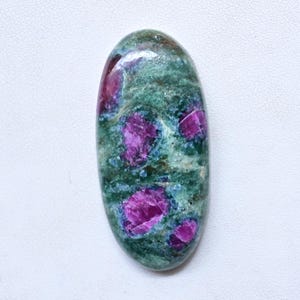 Op de afbeelding: Een ovale edelsteen met een groene basiskleur en levendige magenta insluitsels. De gepolijste steen toont een mix van groene en magenta tinten, met de magenta vlekken verdeeld over het oppervlak. De edelsteen is geplaatst tegen een witte achtergrond.