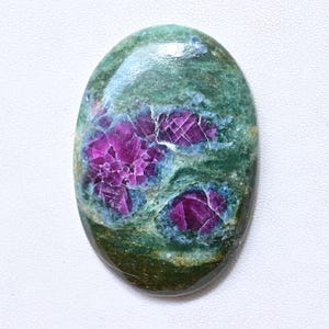 May include: Oval-shaped gemstone with a green base and vibrant magenta inclusions. The stone's surface has a polished, glossy finish. The magenta areas display a fractured, crystalline pattern, adding depth and visual interest to the natural stone.