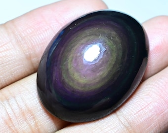 May include: Oval-shaped rainbow obsidian gemstone. The polished stone displays concentric bands of color, including purple, green, and yellow, set against a dark, glossy background. The stone is smooth and reflective, with a high-gloss finish.