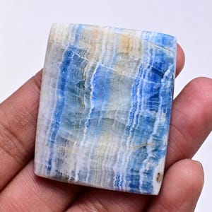 May include: A blue and white banded cabochon gemstone with a square shape. The stone has a layered, striated pattern.