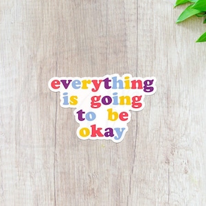 May include: A sticker with the text "everything is going to be okay" in a rainbow of colors on a light wood background.