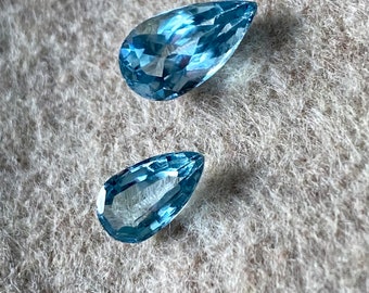May include: Two teardrop-shaped aquamarine gemstones on a light gray fuzzy surface.