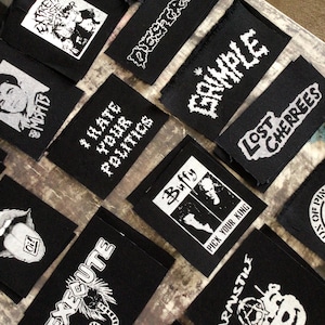Puede incluir: Una colección de parches de tela negra con texto y gráficos blancos. Los parches presentan varios diseños, incluyendo nombres de bandas, lemas e ilustraciones. Algunos de los textos incluyen "Extreme Noise", "I Hate Your Politics", "Buffy", "Pick Your King", "Destroy", "Grimble", "Lost Cherrees", "Execute", "Armistice" y "Oi!"