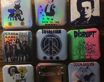 May include: Nine different button badges with various designs. The badges are square and have a silver metal frame. The designs include a peace sign, a cartoon character, a band logo, and a political statement. The badges are all on a wooden surface.