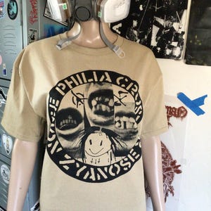 May include: A tan short-sleeved t-shirt with a black and white graphic. The graphic includes the text "PHILIA CR" and "ZYANOSIS" around a drawing of teeth and a smiling face. A respirator mask is worn by the mannequin.