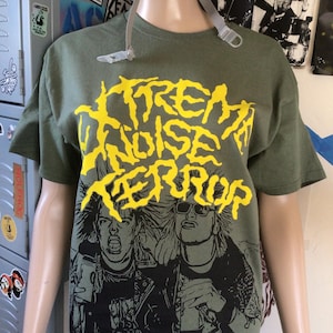Op de afbeelding: Een groen T-shirt met een gele grafische print van de tekst "Extreme Noise Terror" en een zwart-witte illustratie van twee mensen.