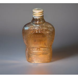 Könnte beinhalten: Eine Vintage-Glasflasche in Bernsteinfarbe mit dekorativem Design und Metallkappe. Die Flasche hat eine einzigartige Form und trägt den Text "SINCE 1880". Die Flasche ist leer und scheint in gutem Zustand zu sein.