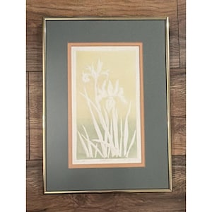 May include: Framed art print featuring a botanical illustration of white iris flowers and leaves against a gradient yellow and green background. The artwork is set within a peach-colored border, a gray mat, and a gold-tone frame.