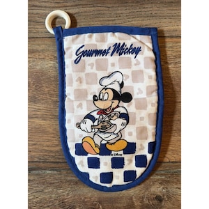 Puede incluir: Un guante de cocina blanco con borde azul y una anilla para colgar. El guante presenta un gráfico de Mickey Mouse con gorro y delantal de chef mientras sostiene una cuchara de madera. El texto Gourmet Mickey está impreso en azul.