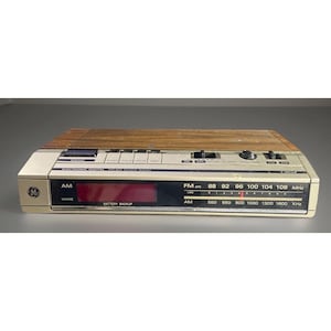 Puede incluir: Un radio reloj digital AM/FM GE vintage con una parte superior con vetas de madera y una parte frontal de color crema. La radio cuenta con una pantalla LED roja, un dial de frecuencia AM/FM y varios botones de control. El radio reloj tiene una función de respaldo de batería.