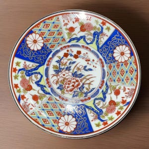 Könnte beinhalten: Ein runder, dekorativer Teller mit weißem Grund und Goldrand. Der Teller zeigt ein zentrales Blumendesign, das von blau, rot und gelb gemusterten Abschnitten umgeben ist. Das Design umfasst Blumen, geometrische Formen und bandartige Elemente.