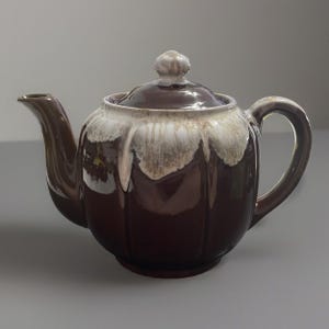 May include: A brown ceramic teapot with a lid and handle. The teapot has a drip glaze effect in shades of white and brown. The spout and handle are a darker brown.