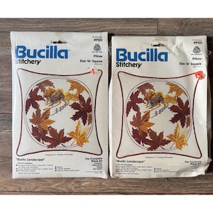May include: Two Bucilla Stitchery pillow kits. Each kit features a 16-inch square pillow design with a rustic landscape scene surrounded by autumn leaves in shades of brown, orange, and yellow. The kits include the complete pillow kit with a zipper.