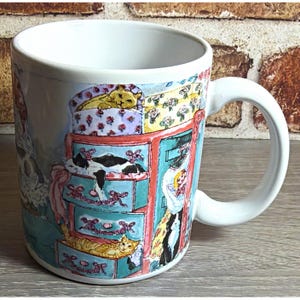 May include: A white ceramic mug featuring a colorful watercolor-style illustration of several cats resting in and on a multi-drawer dresser. The artwork depicts cats in various poses, including one in a top drawer and one on the dresser top.